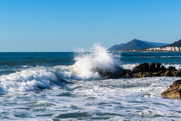 A wave crashes against the rocks on a sunny day at Vila Praia de Âncora, Portugal. Stunning coastal landscape with blue sky, white foam, and distant mountains in the background.