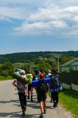 Obraz premium backs of guys, sleeping bags, backpacks, go, summer hats, country road, village, private houses, street, warmth, green leaves, trees, teenagers, forest, travel, Europe, Ukraine, background blue sky, e