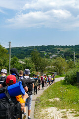 backs of guys, sleeping bags, backpacks, go, summer hats, country road, village, private houses, street, warmth, green leaves, trees, teenagers, forest, travel, Europe, Ukraine, background blue sky, e