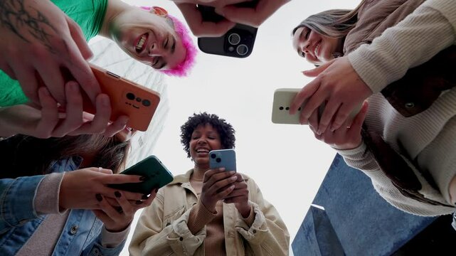 Low angle view of young group of people using mobile phones together at city street. Diverse college student friends watching social media content on cell. Cellphone addicted people concept. - Powered by Adobe