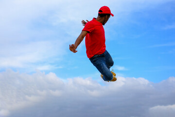 A man is jumping in the air with his arms outstretched. The sky is blue and there are clouds in the background. Scene is joyful and energetic