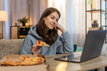 Happy young Caucasian woman eating delicious junk food pizza sitting on sofa at table watching...