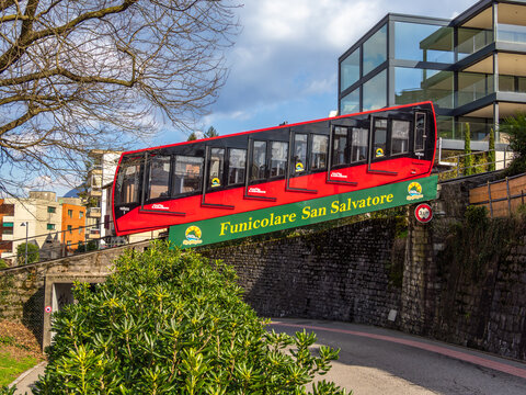 Lugano, Switzerland - March 26, 2025: A red funicular train San Salvatore ascending a steep track, modern buildings in the background.