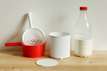 Plastic equipment for making soft cheese and cottage cheese at home. A white uniform, a red ladle and a bottle of milk.