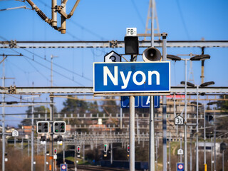 A blue and white train station sign displaying the name NYON against a backdrop of railway...