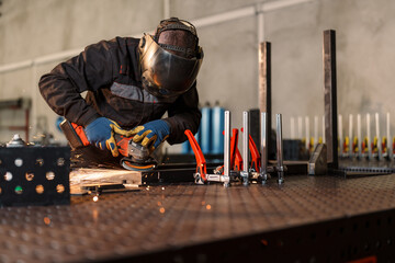 Metalworker wearing protective gear uses an angle grinder, creating sparks while working on a metal piece clamped to a workbench in a workshop