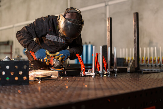 Metalworker wearing protective gear uses an angle grinder, creating sparks while working on a metal piece clamped to a workbench in a workshop