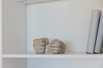 A modern white bookshelf featuring unique concrete hand sculptures and minimalist decor elements in a stylish home interior. © svittlana