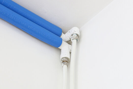 Insulated water pipes system. Blue foam thermal cover. White plumbing tubes in corner. Central heating installation view. Indoor pipe connection detail. Modern home energy saving solution.