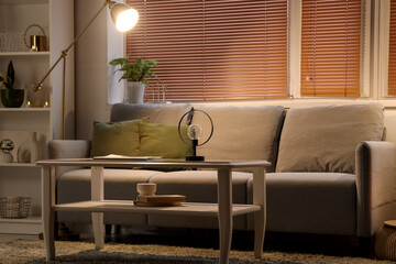 Interior of modern living room with grey sofa, coffee table and glowing lamp at evening