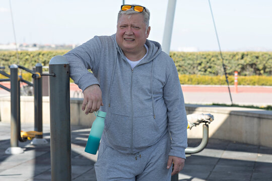 an older man  taking a break at an outdoor fitness park, holding a turquoise water bottle in his right hand. The background includes outdoor gym equipment