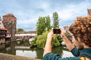 Nuremberg,Germany, August 1, 2023. A young Caucasian man is fascinated by the enchanting view of the river with the half-timbered house reflected in the water. He uses his cell phone to photograph it.