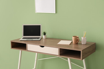 Workplace with laptop, houseplant, cup and stationery near green wall in office