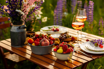Elegant gorgeous wedding table decor or romantic dinner arrangement outdoors in blooming field. Purple lupine flowers, candles, fruits and wine, wooden vintage furniture. Sunset, summer, golden hour