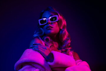 Elegant young woman in glamorous neon lighting wearing stylish sunglasses and luxurious coat.