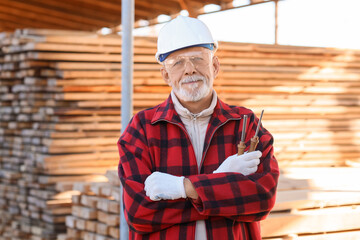 Happy mature male carpenter in hardhat with professional tools at sawmill