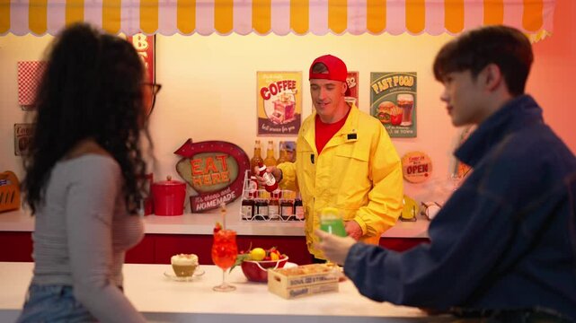 Happy man with yellow shirt turn around to pick and add tabasco sauce in to food while standing behind bar. Attractive diverse friend talking and enjoy eating while holding glass of juice. Regalement.