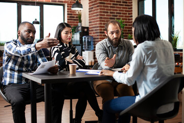 Black female applicant seated in job interview, serious diverse recruiters asking questions across table in modern office. Asian woman with arms crossed and men gesturing to lady and questioning her.