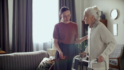 Young woman gently helping elderly grandmother with walker in cozy living room, providing mobility assistance during day at home - Powered by Adobe