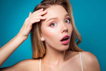 Obraz premium Young blonde woman showing a surprised expression while touching her hair against a blue background