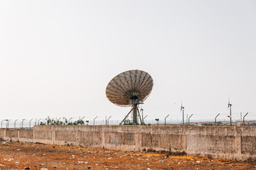 A large satellite dish is sitting on a hillside next to a windmill. The scene is peaceful and...