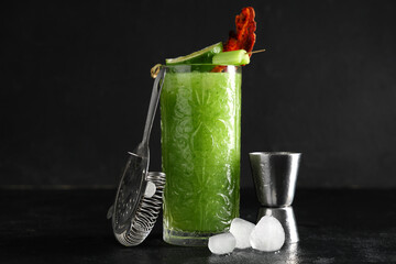 Strainer, ice cubes, glass of bloody Mary cocktail garnished with bacon and celery on black...