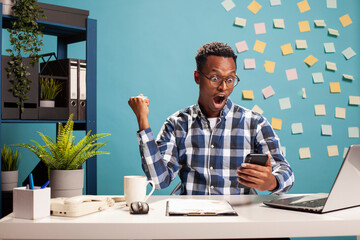 Excited male entrepreneur holding smartphone and celebrating big win in online video game. Black...