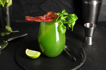 Glass of green bloody mary cocktail with bacon, celery and lime on black background, closeup
