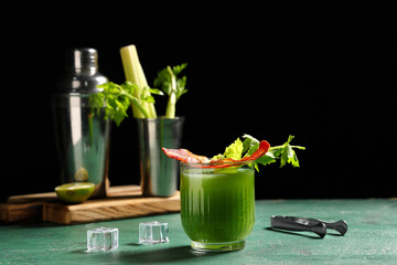 Glass of green bloody mary cocktail with bacon, celery and ice cubes on color table against black...