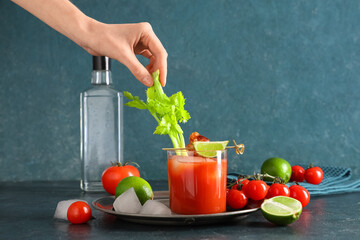 Female hand with celery and glass of bloody mary cocktail with bacon on blue background