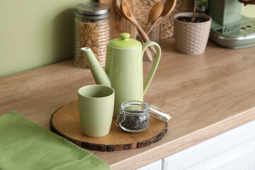 Teapot with jar of dry tea and cup on table near green wall in kitchen
