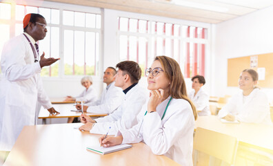 Interested young latin american woman in white coat listening to lecture and taking notes in...
