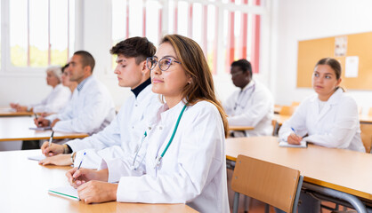 Latin young woman medical student in white coat attedning classes in university, sitting at desk and listening to lecture.
