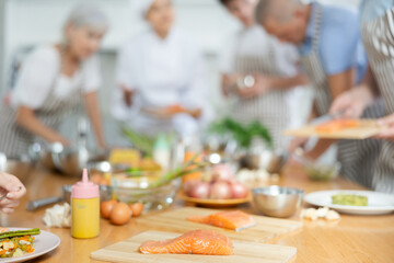 Background cook leads master class in cooking in kitchen. In foreground, piece of raw salmon fillet...
