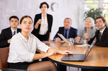Portrait of positive young female office worker sitting at table with colleagues, listening with...