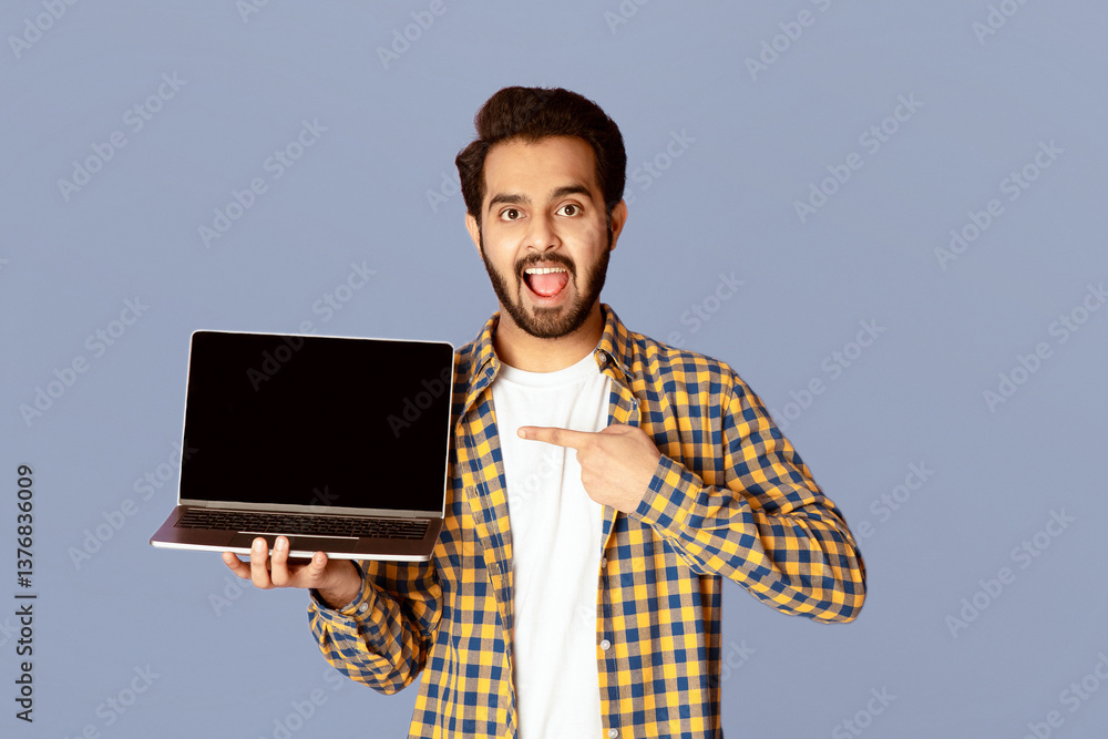 Poster excited indian guy pointing at empty laptop screen over violet background, mockup for website design