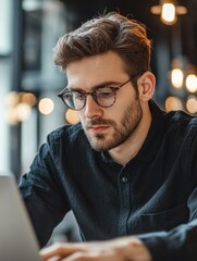 Obraz premium An adult male focused on a laptop screen, wearing glasses and black shirt, indoors during the day.