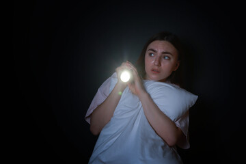 Fear of darkness. Scared young woman with flashlight hugging pillow on black background, space for...