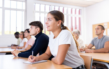 Interested motivated girl listening with attention and taking notes of lecture while studying in university classroom among groupmates