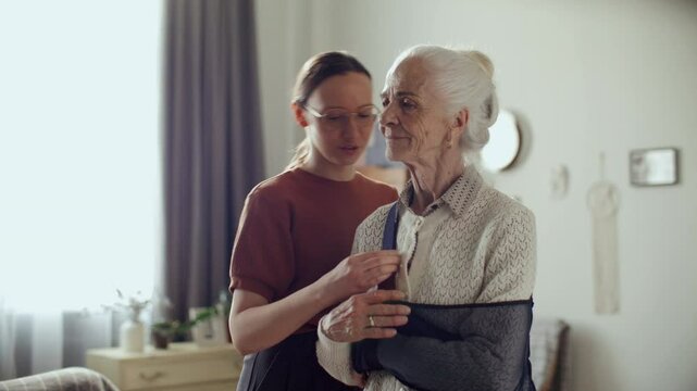 Young woman talking to elderly grandmother as adjusting medical arm sling in living room, assisting her during recovery at home