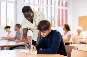 Strict African American teacher standing next to focused young guy sitting at desk during lesson, pointing out error in solving task in student notebook..