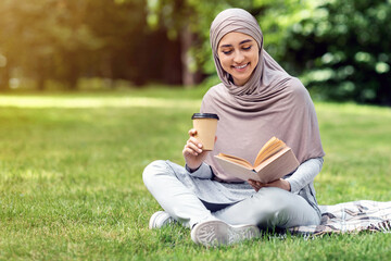 Pretty arab girl reading book and drinking coffee or tea at park, sitting on grass