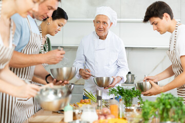 Elderly man cook in uniform teaches group of different people how to cook dish
