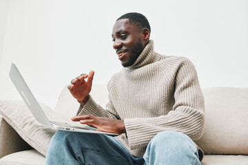 Young Black man in a cozy sweater smiles while using a laptop at home, showcasing a relaxed...