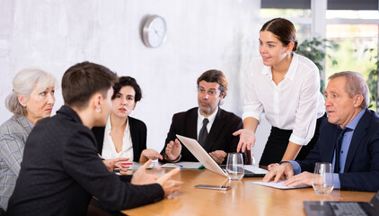 Successful smiling young businesswoman explaining new business strategy to partners sitting at table in office