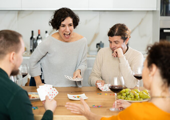 Cheerful friendly company of men and women sitting at kitchen table and enjoying the poker game. Friends play a card game at home. Evening entertainment concept