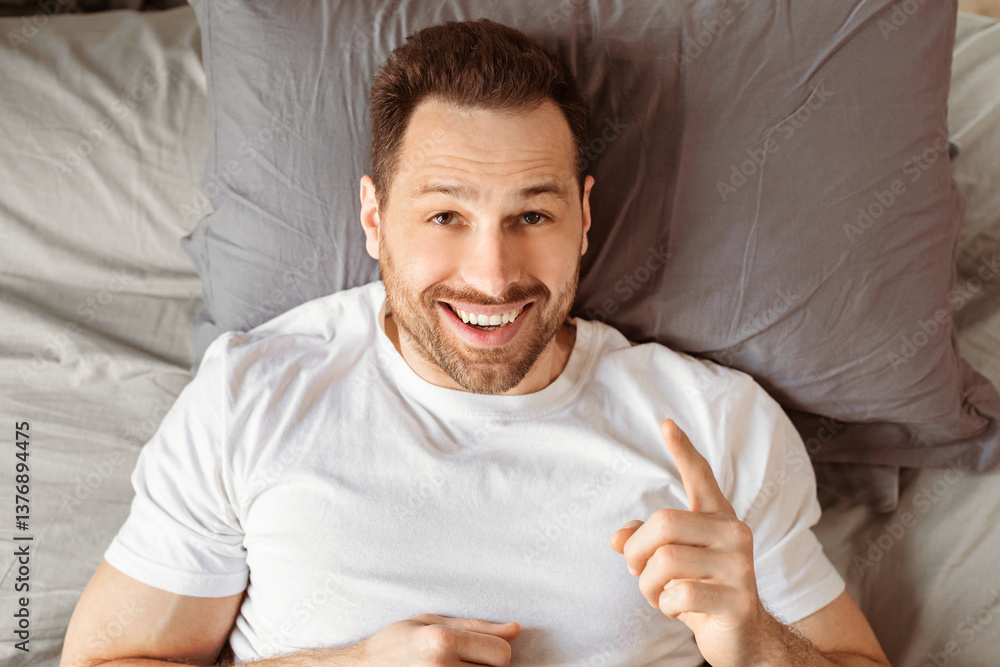 Poster joyful guy having idea pointing finger up lying in bed, posing looking at camera in bedroom at home.