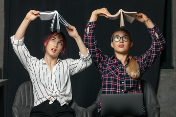 Two young women sitting in a modern office, working together on a laptop. One points at the screen...
