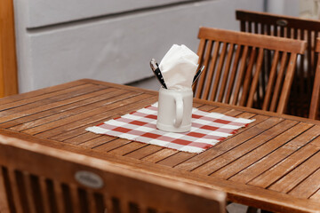 A charming rustic outdoor dining setup featuring a wooden table with a checkered placemat, a ceramic mug holding napkins and cutlery, creating a cozy and inviting atmosphere perfect for al fresco