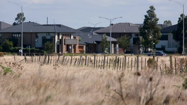 Dry grasses sway with a rustic fence line dividing the undeveloped land from a modern Australian suburban housing neighborhood in an outer suburb of Melbourne.Concept of drought and urban fringe